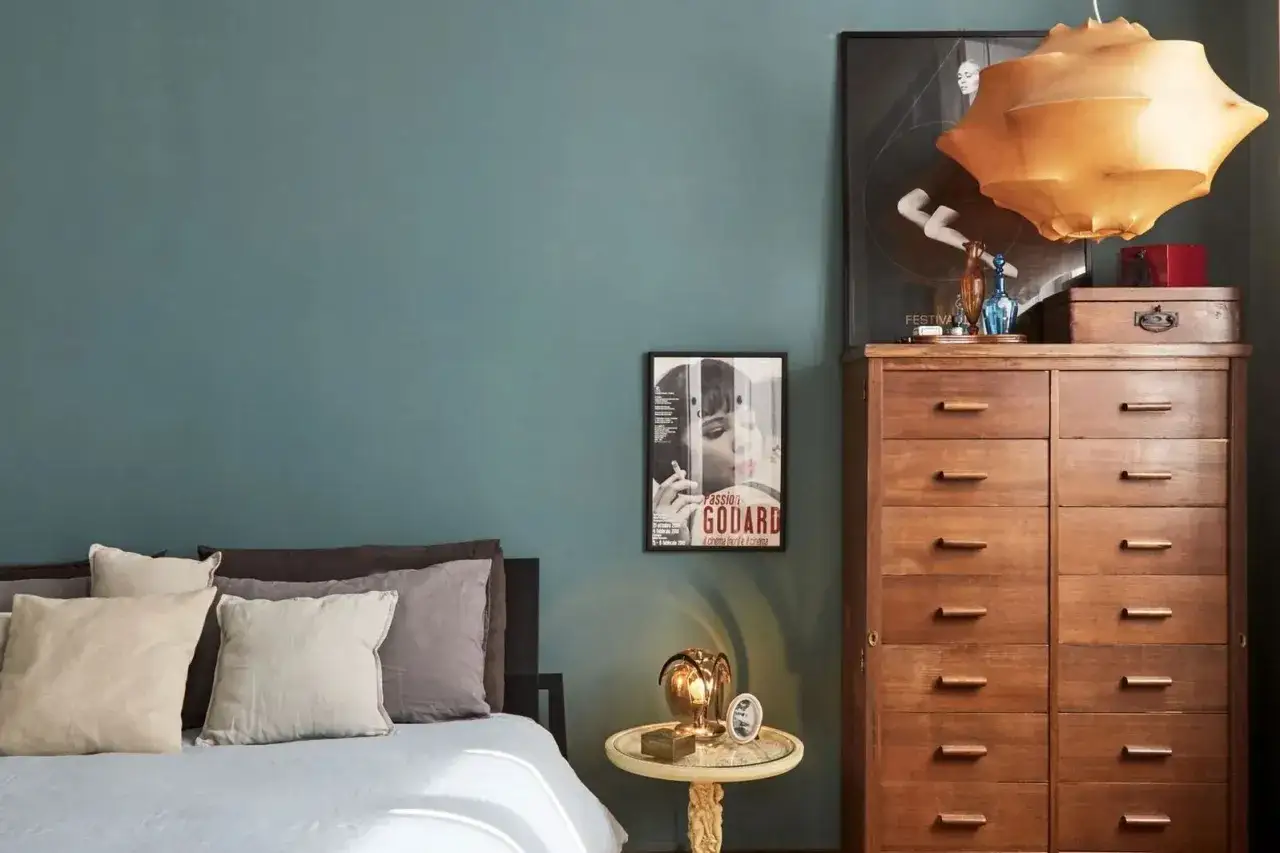 Camera da letto con pareti color petrolio, un invito alla cromoterapia. Letto con cuscini morbidi, comodino con lampada vintage e un mobile in legno scuro.