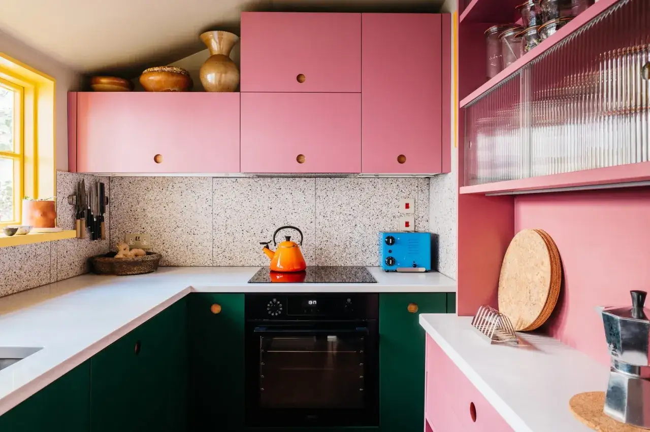 Cucina rosa con mobili verdi, piano cottura nero, tostapane blu e bollitore arancione. Pareti a mosaico e decorazioni in ceramica.