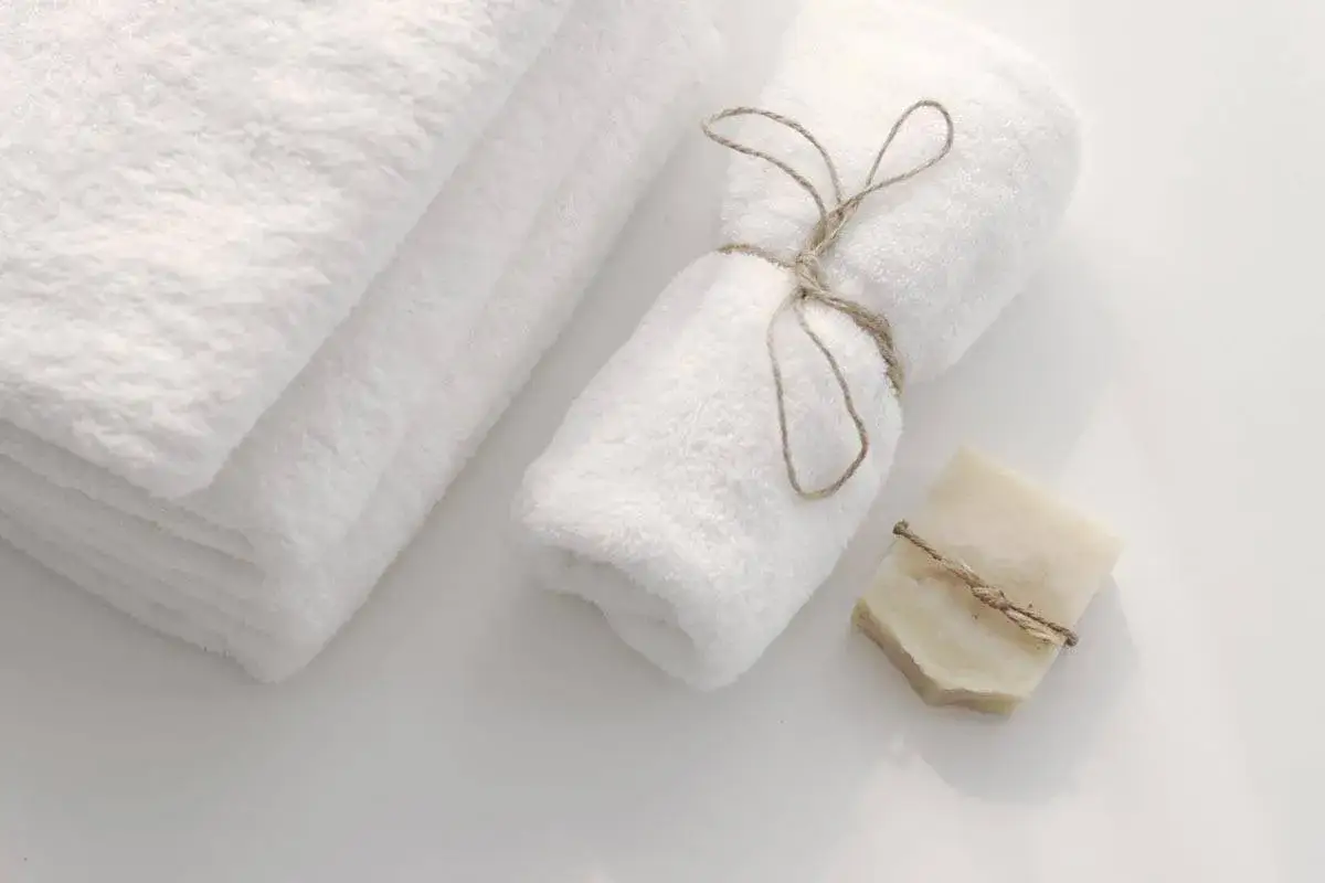 A stack of fluffy white towels and one rolled and tied with twine, next to a bar of soap. Elegante modo per piegare gli asciugamani.