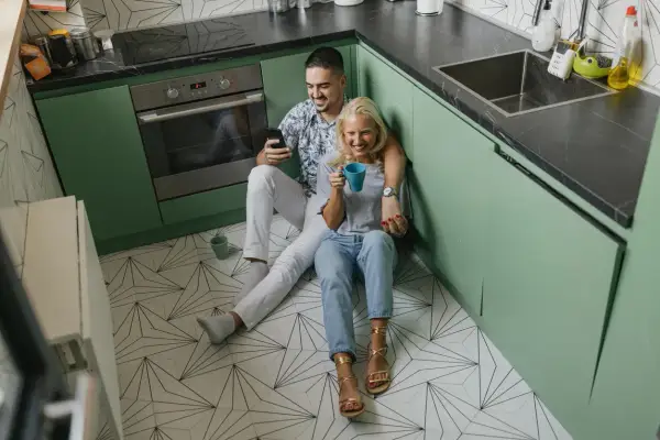 Coppia rilassata in cucina, con mobili verde menta e un pavimento geometrico. Lui guarda il telefono, lei sorseggia una bevanda.