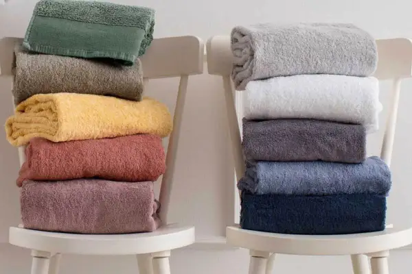 Ecco come piegare gli asciugamani in modo elegante: pile di asciugamani colorati e neutri disposti su sedie bianche.