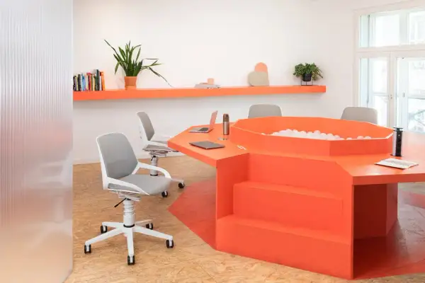 Un ufficio moderno con un tavolo arancione a forma di esagono, dotato di una vasca di palline e sedie ergonomiche.
