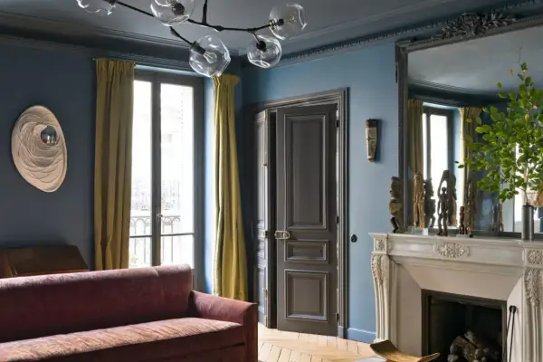 Soggiorno elegante con divano bordeaux, pareti blu balena e tendaggi dorati. Un camino in marmo e sculture africane completano l'arredamento, creando abbinamenti sofisticati.