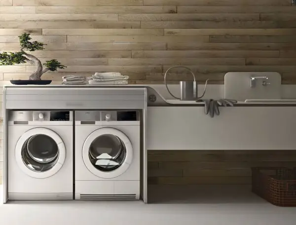 Lavatrice in cucina moderna, con due elettrodomestici bianchi affiancati sotto un piano di lavoro con bonsai e asciugamani.