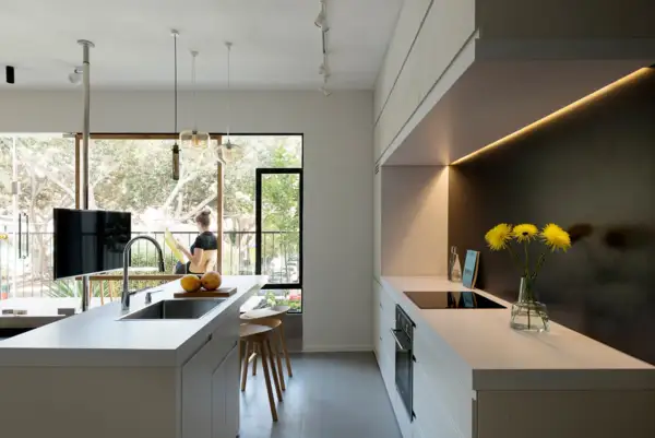 Un moderno cucina soggiorno open space di 50 mq con isola centrale, bancone con lavello e un'area cottura lineare. Una donna legge vicino alla finestra.