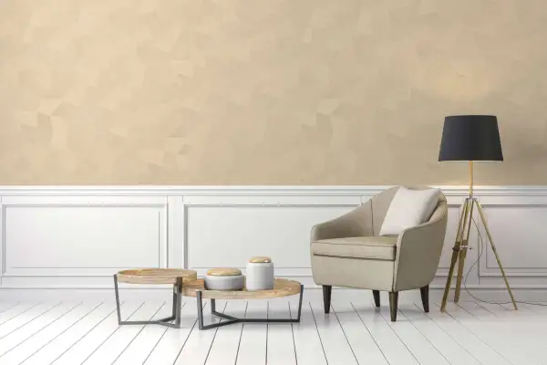 Poltrona beige e tavolini in legno su pavimento bianco, con parete in color champagne e lampada nera.