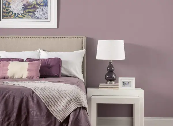 Letto con testiera imbottita, cuscini viola e una coperta color malva. Sul comodino, una lampada e una cornice con una farfalla.