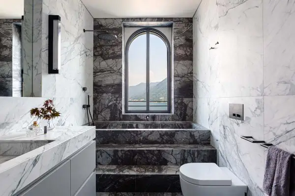 Bagno di lusso con doccia in muratura, vasca e lavabo in marmo, affacciato su un lago e montagne.