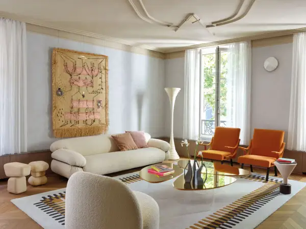Soggiorno moderno con divano bianco, poltrone arancioni e arazzo colorato appeso alle pareti color crema.